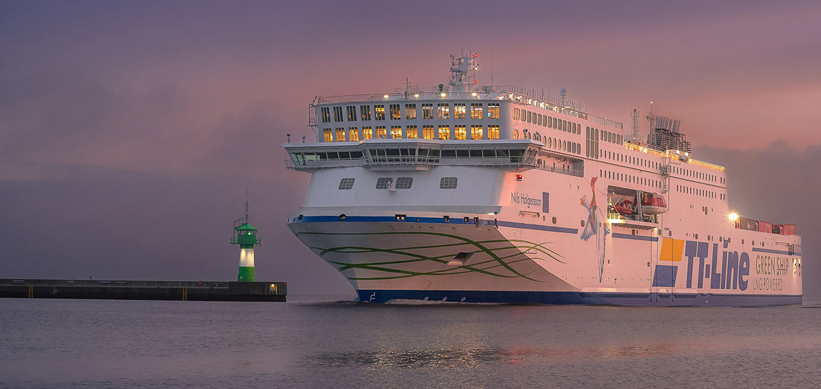 Ein großes Fährschiff der TT-Line fährt bei Sonnenuntergang in einen Hafen ein. Im Hintergrund ist ein grüner Leuchtturm zu sehen.