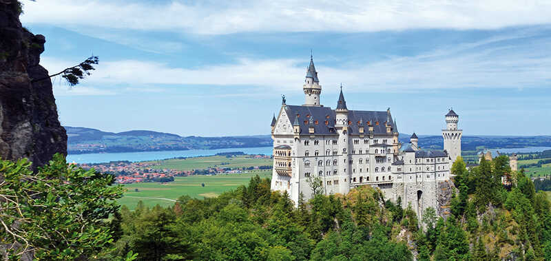 Das Bild zeigt ein märchenhaftes Schloss mit zahlreichen Türmen, das auf einem bewaldeten Hügel vor einer weiten Landschaft liegt.