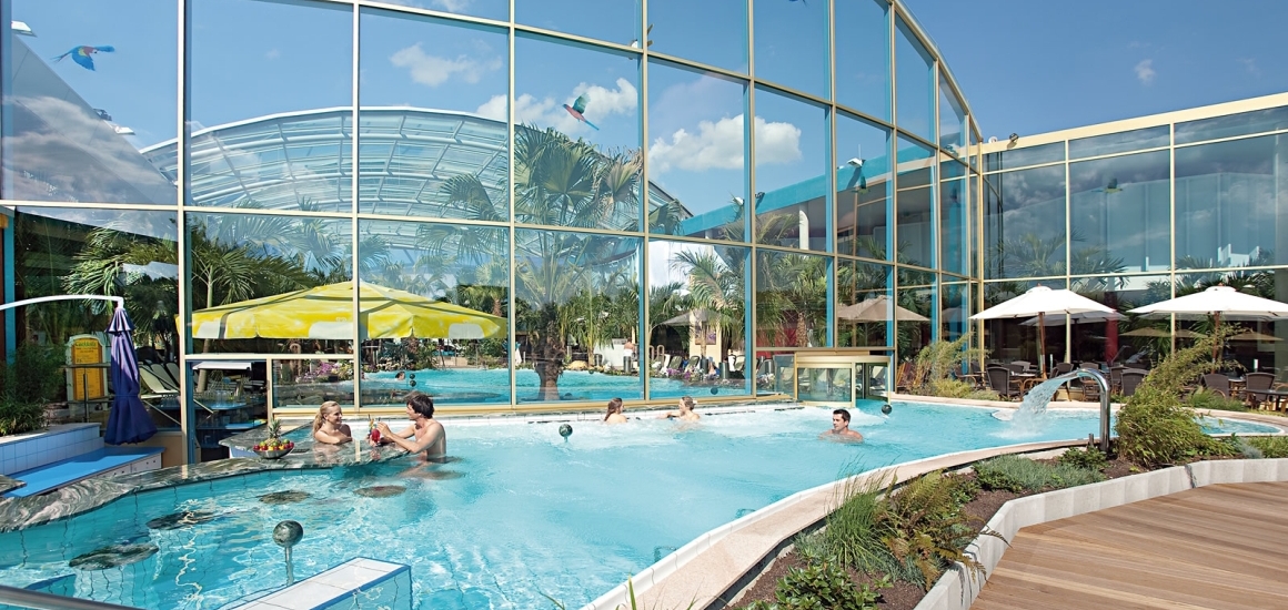 Außenansicht einer Therme mit Pool, Glasfassade und Sonnenschirmen