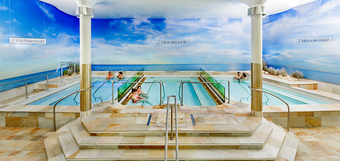 Innenansicht einer Therme mit mehreren Becken und Menschen, die sich entspannen. Die Wände sind mit einem Wandbild eines blauen Himmels und Meeres dekoriert.