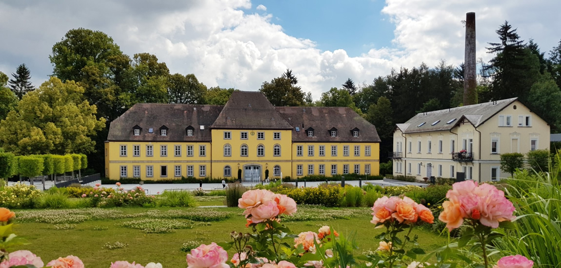 Ein gelbes Schlossgebäude mit einem großen Garten im Vordergrund, umgeben von Bäumen und einem blauen Himmel mit Wolken.