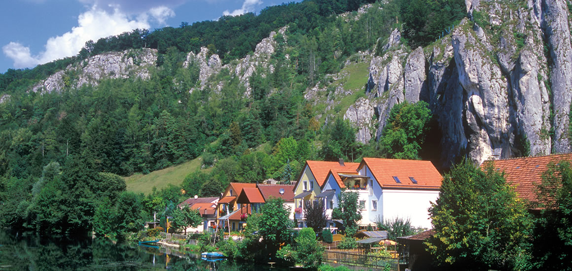 Landschaftsbild der Altmühl in Bayern mit Fluss, Häusern und Felsen im Hintergrund.