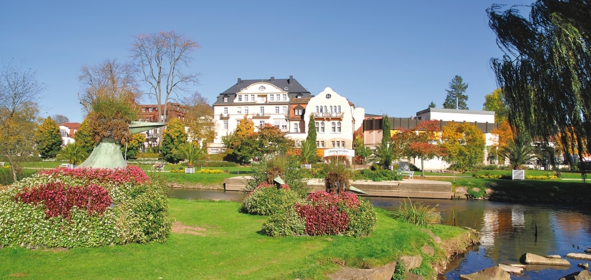 Kurpark in Bad Kissingen mit gepflegten Grünflächen, Blumenbeeten und einem Fluss im Vordergrund, umgeben von historischen Gebäuden.