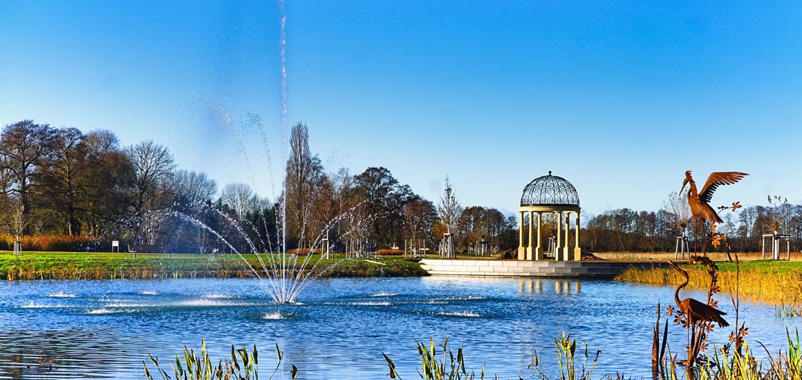 Ein Teich mit einer Fontäne in der Mitte, umgeben von Bäumen und einem Pavillon im Hintergrund. Im Vordergrund ist eine Skulptur eines Vogels zu sehen.