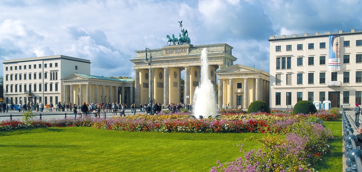 Brandenburger Tor mit einer Fontäne im Vordergrund und blühenden Gärten, umgeben von historischen Gebäuden.