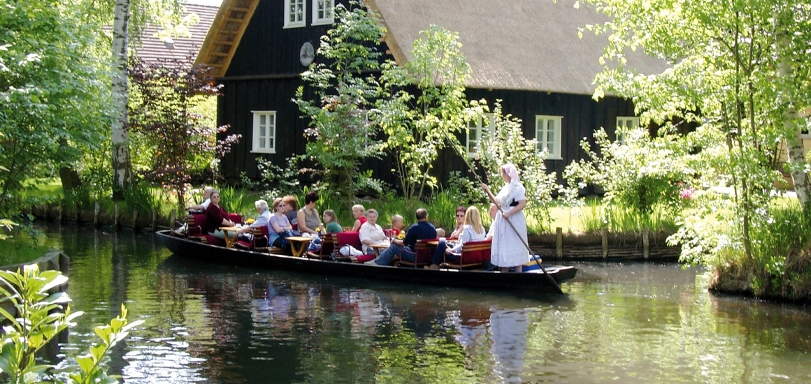 Eine Gruppe von Menschen sitzt in einem traditionellen Kahn, der von einer Person mit einem langen Stab durch einen ruhigen Wasserkanal im Spreewald gesteuert wird. Im Hintergrund ist ein traditionelles Haus mit Reetdach zu sehen, umgeben von üppiger Vegetation.