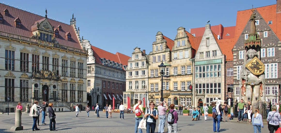 Marktplatz in Wittmund, Deutschland mit Bronzestatue eines sitzenden Mannes und Schafen auf einem gepflasterten Platz, umgeben von historischen Backsteingebäuden.