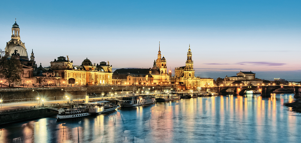 Abendansicht der Dresdner Altstadt mit beleuchteten historischen Gebäuden und der Elbe im Vordergrund.