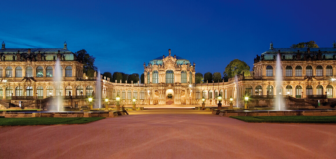 Nachtaufnahme des Dresdner Zwingers mit beleuchteter Architektur und Springbrunnen im Vordergrund.