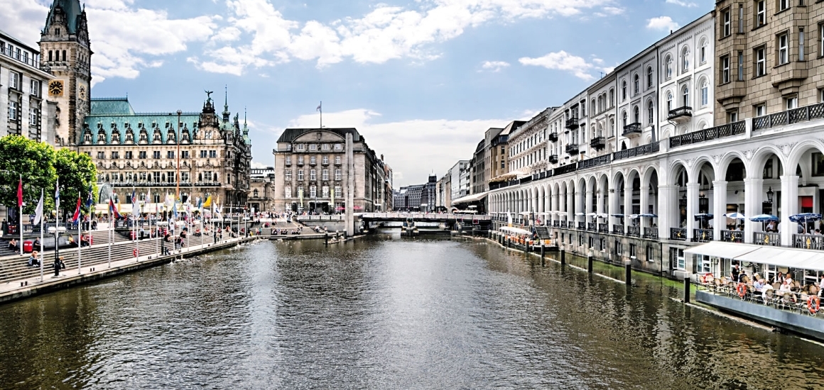 Blick auf die Alsterarkaden in Hamburg mit Wasserkanal im Vordergrund und historischen Gebäuden mit Arkaden im Hintergrund.