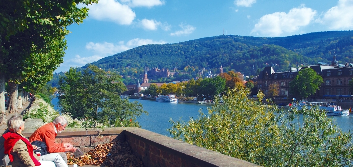 A panoramic view of Heidelberg, showcasing a couple overlooking the city and the Neckar River, with a backdrop of red-roofed buildings and green hills.