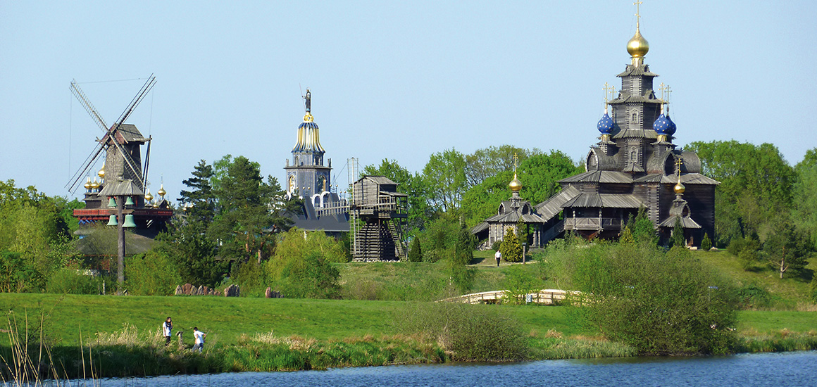 Eine Landschaft mit einer Windmühle, mehreren Gebäuden mit Zwiebeltürmen und einem See im Vordergrund, umgeben von Bäumen und Wiesen.