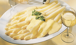 Ein Teller mit weißen Spargelstangen, bedeckt mit Sauce Hollandaise, dekoriert mit Kräutern. Daneben steht ein Glas Weißwein.
