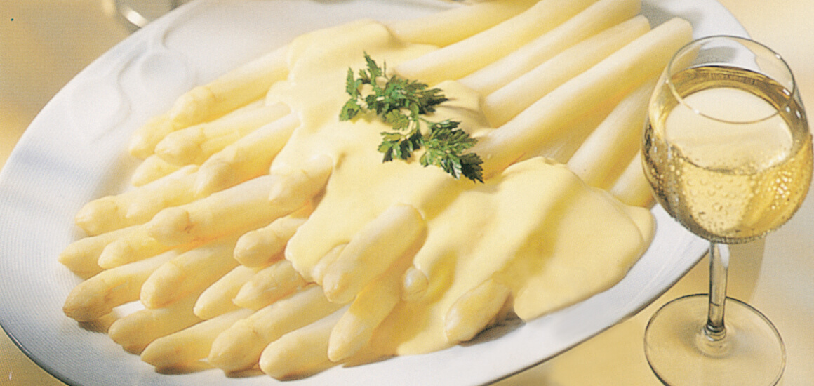 Ein Teller mit weißem Spargel, bedeckt mit Sauce Hollandaise, daneben ein Glas Weißwein.