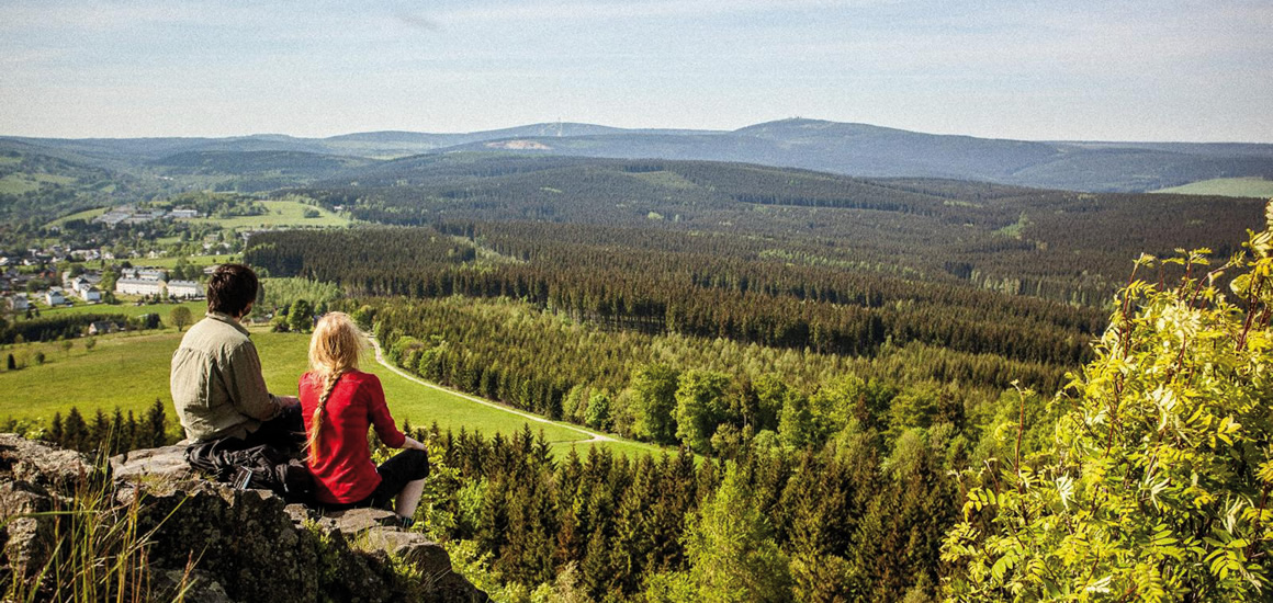 Zwei Personen sitzen auf einem Felsen und blicken auf eine weitläufige Waldlandschaft im Erzgebirge.