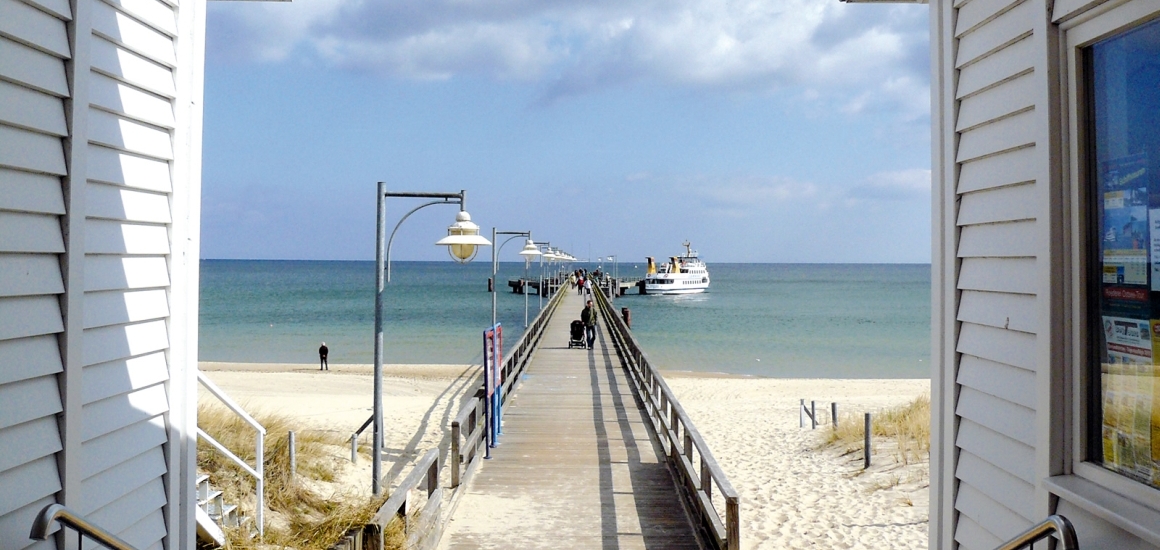 Ein langer Holzsteg führt über den Sandstrand zu einem Anlegesteg im Meer, an dem ein Schiff festgemacht ist. Der Himmel ist leicht bewölkt, und es sind einige Menschen auf dem Steg zu sehen.