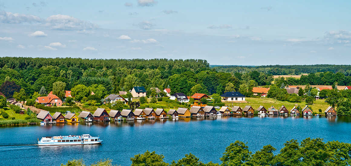 Das Bild zeigt einen idyllischen See mit einem Ausflugsschiff, bunten Bootshäusern am Ufer und viel grüner Natur im Hintergrund.