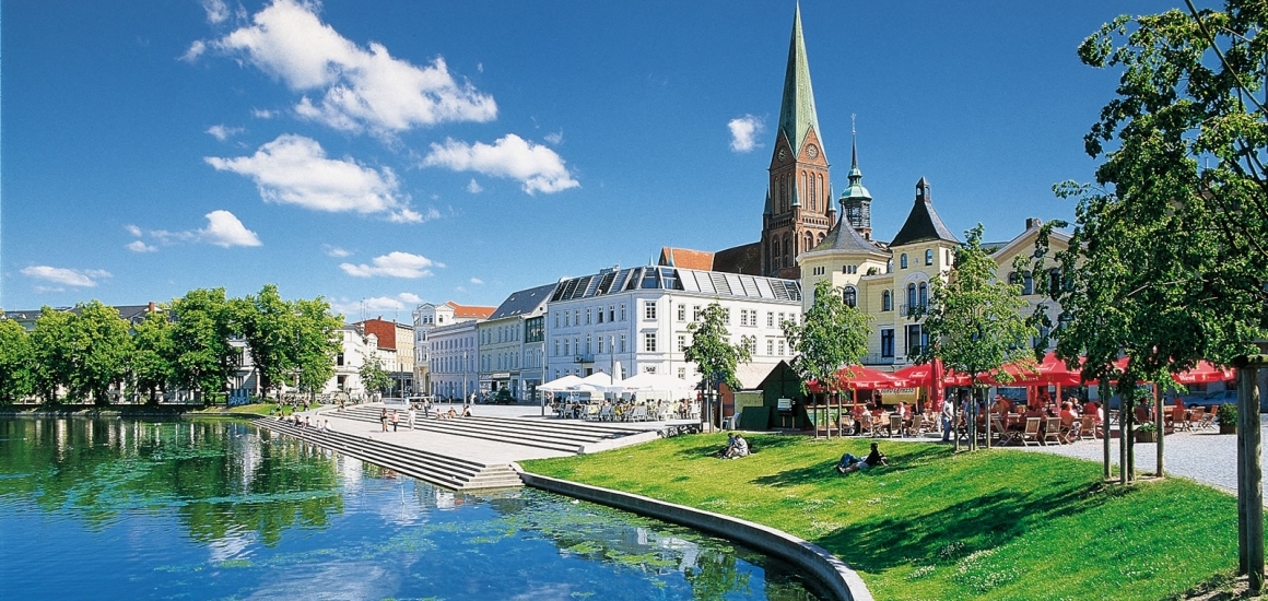 Blick auf den Pfaffenteich in Schwerin mit umliegenden Gebäuden und der Schweriner Dom im Hintergrund.