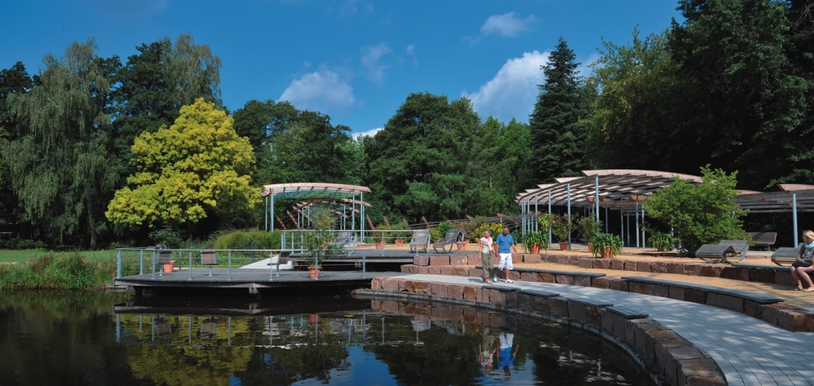 Ein Park in Bad Bevensen, Niedersachsen, mit einem Teich, umgeben von Bäumen und Sitzbereichen. Menschen spazieren entlang eines gepflasterten Weges.