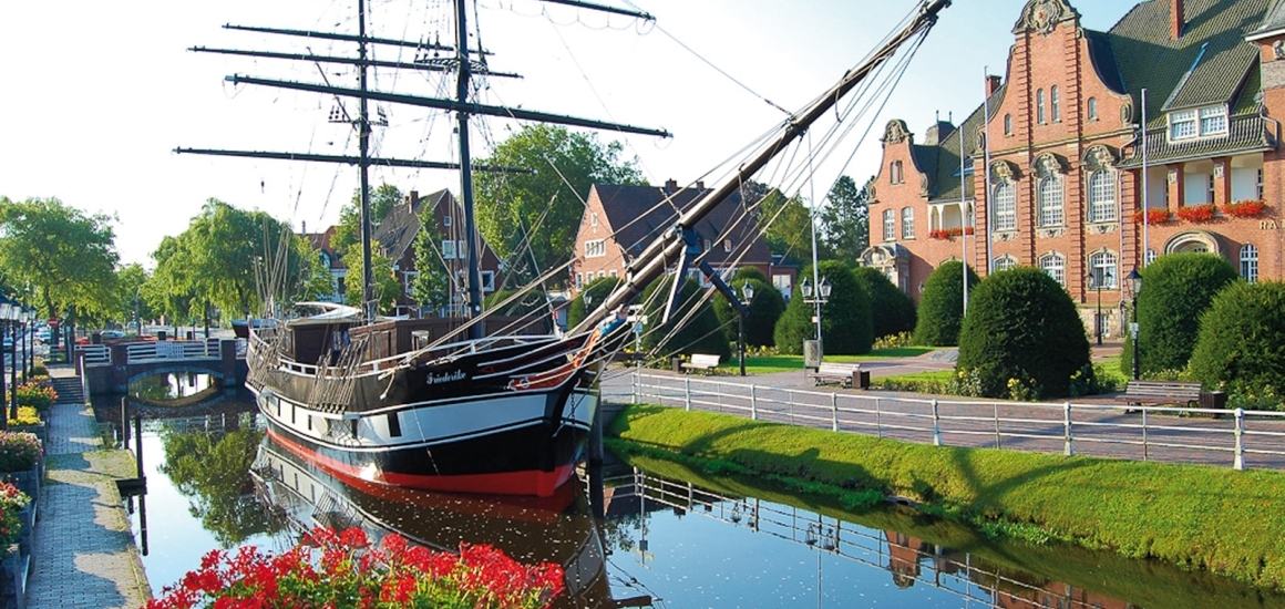 Ein historisches Segelschiff liegt in einem Kanal in Papenburg, umgeben von gepflegten Grünflächen und historischen Gebäuden.