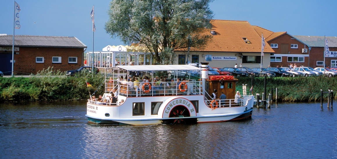 Ein Fluss mit einem kleinen Ausflugsboot, das den Namen 'Concordia II' trägt. Im Hintergrund sind Gebäude und geparkte Autos zu sehen. Die Szene wirkt ruhig und sonnig.