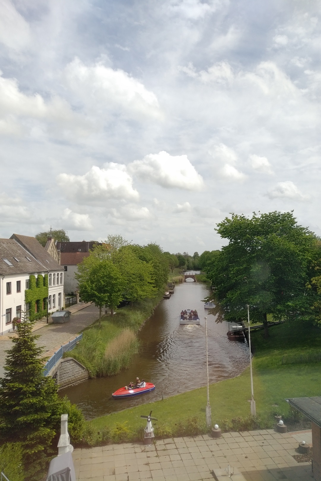 Ein Fluss mit einem kleinen Boot und einem größeren Boot im Hintergrund, umgeben von Bäumen und Gebäuden unter einem bewölkten Himmel.