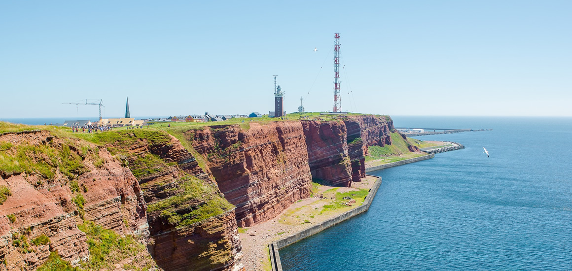 Küstenlandschaft von Helgoland mit roten Klippen und Funkturm, umgeben von blauem Meer und klarem Himmel.