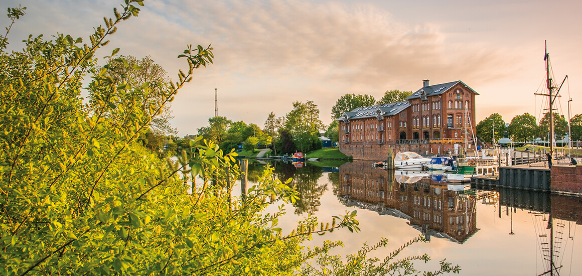 Ein malerisches Bild eines Hafens bei Sonnenuntergang mit einem Backsteingebäude am Wasser, umgeben von Bäumen und Booten.