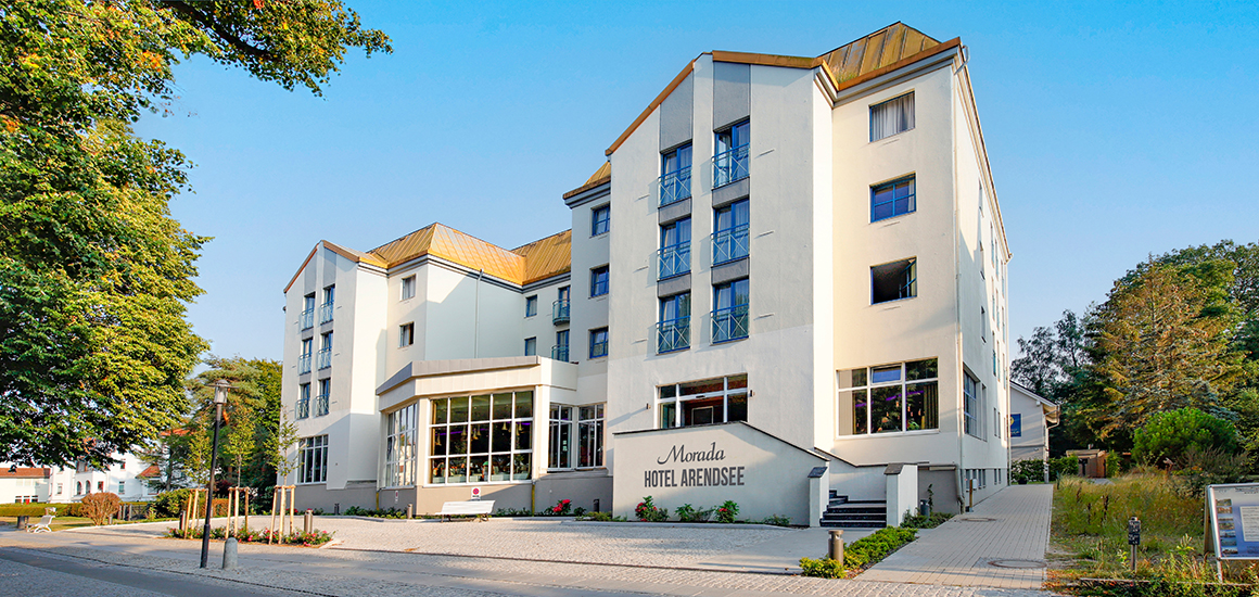 Außenansicht eines modernen Hotels mit mehreren Stockwerken und großen Fenstern, umgeben von Bäumen und einem klaren Himmel.