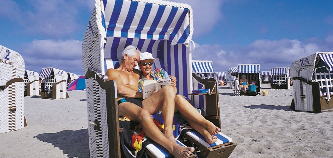 Ein älteres Paar sitzt in einem Strandkorb am Strand von Kühlungsborn, umgeben von weiteren Strandkörben. Sie wirken entspannt und genießen den sonnigen Tag.