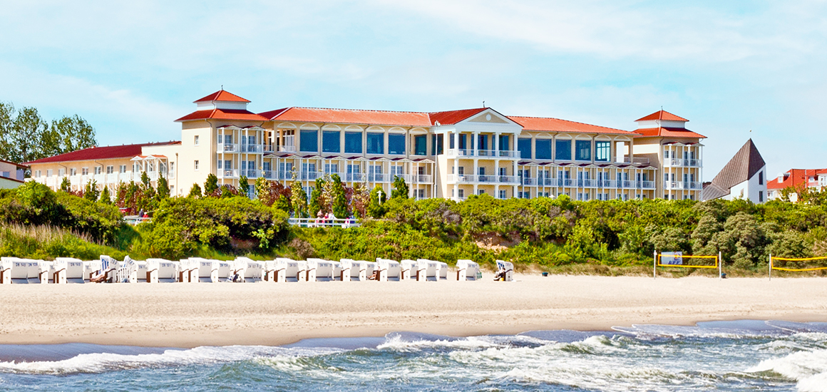 Ansicht eines großen Hotels mit rotem Dach und vielen Fenstern, gelegen an einem Strand mit Strandkörben und grünem Bewuchs im Vordergrund.