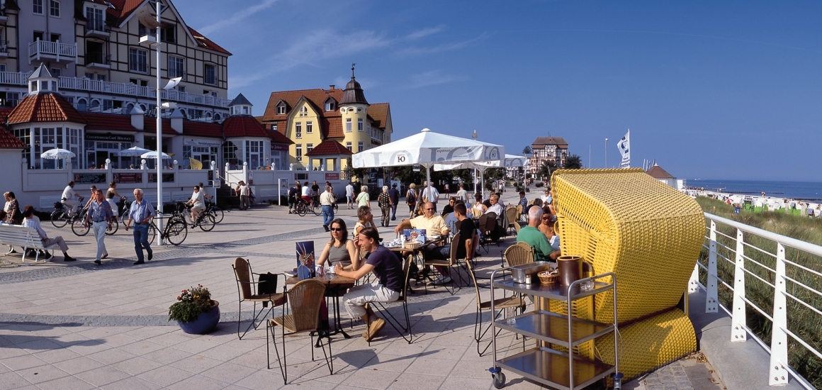 Menschen sitzen an Tischen auf einer Promenade in Kühlungsborn, Ostsee, mit Gebäuden im Hintergrund und einem Strandkorb im Vordergrund.