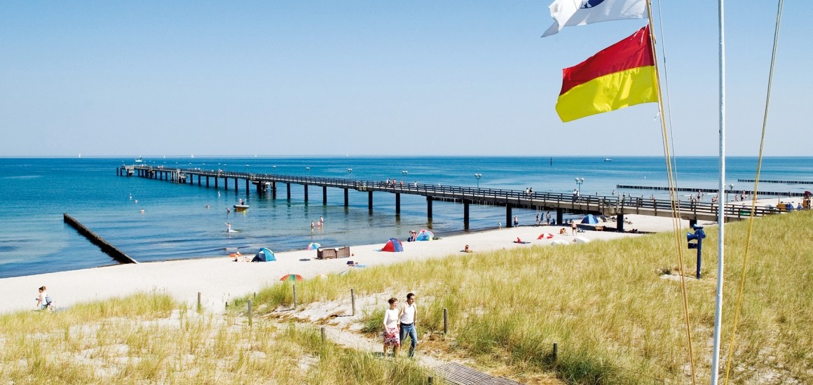 Seebrücke in Graal-Müritz an der Ostsee mit Strand und wehenden Flaggen im Vordergrund.