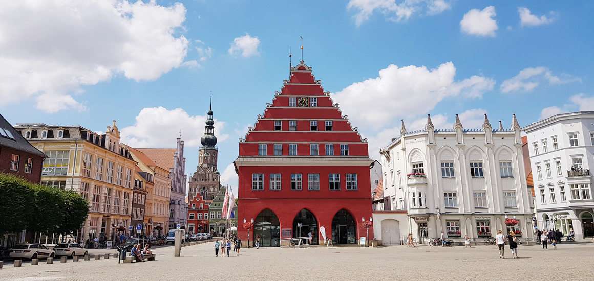 Historisches Gebäude am Marktplatz von Greifswald mit roter Fassade und Treppengiebel, umgeben von weiteren historischen Gebäuden und Menschen auf dem Platz.