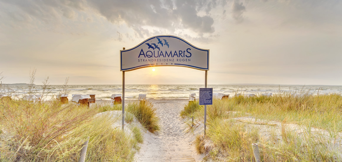 Strandzugang in Juliusruh an der Ostsee mit einem Schild 'Aquamaris' und Strandkörben im Hintergrund.