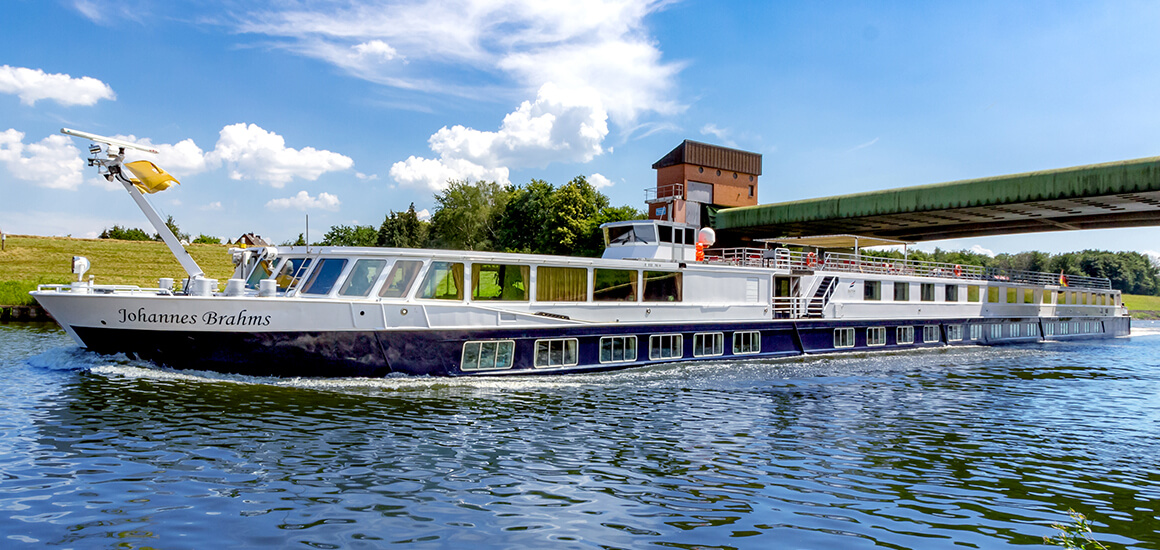 Ein Flusskreuzfahrtschiff namens 'Johannes Brahms' fährt auf einem Fluss unter einer Brücke hindurch. Das Schiff hat mehrere Decks und ist von Bäumen und einem blauen Himmel umgeben.
