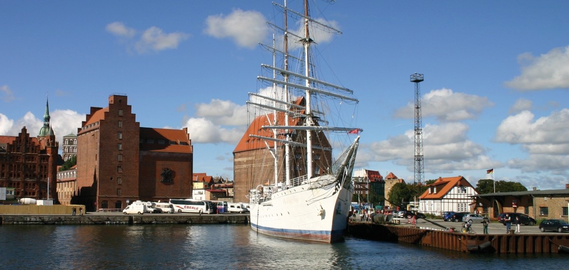 Ein Hafen in Stralsund mit einem großen Segelschiff und historischen Gebäuden im Hintergrund.