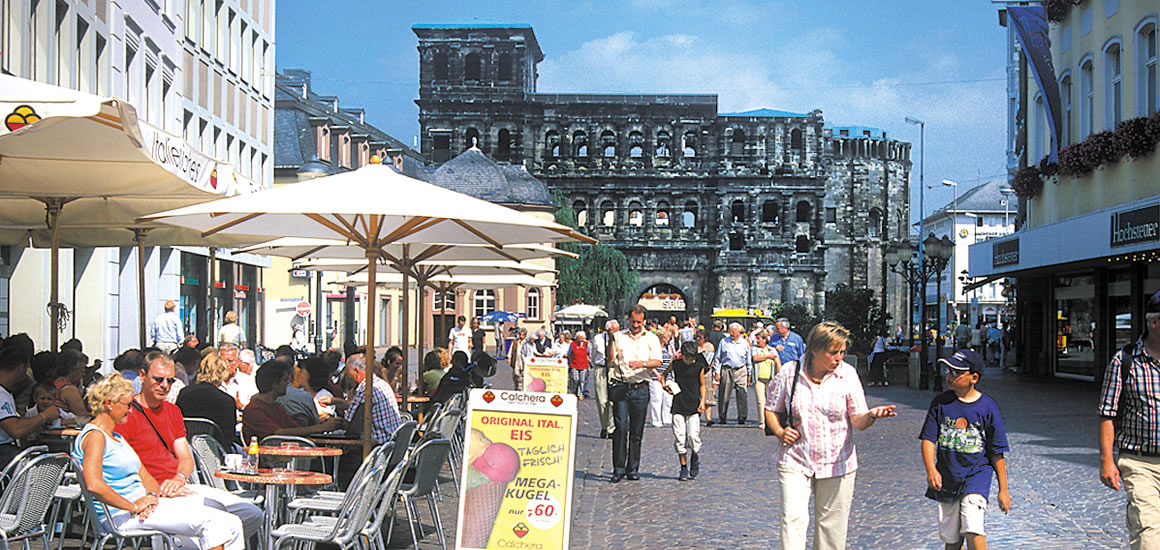 Straßenszene in Trier mit der Porta Nigra im Hintergrund, Menschen sitzen in einem Straßencafé und gehen die Straße entlang.