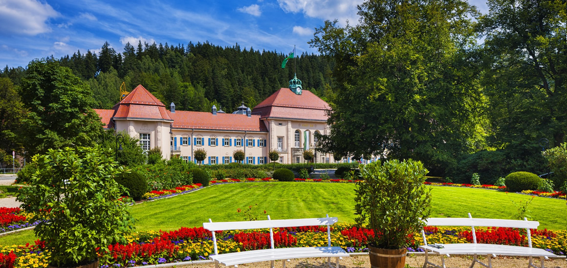 Ein gepflegter Park mit Blumenbeeten und Bänken vor einem historischen Gebäude in Bad Elster, Sachsen.