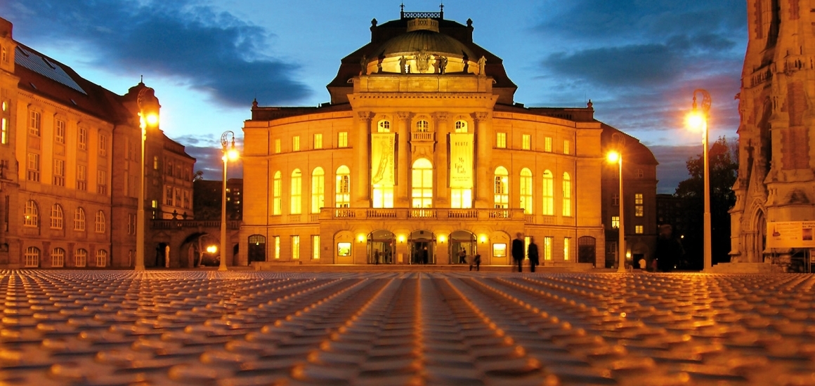 Beleuchtetes Opernhaus in Chemnitz bei Dämmerung, mit gepflastertem Vorplatz und umgebenden Gebäuden.