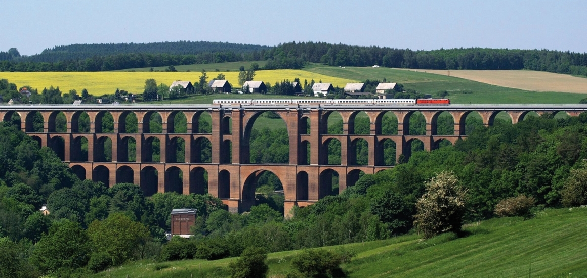 Göltzschtalbrücke in Sachsen, Deutschland, mit einem Zug, der darüber fährt. Die Brücke ist von grüner Landschaft und einigen Häusern umgeben.
