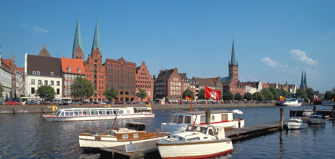 Blick auf die Altstadt von Lübeck mit der Trave im Vordergrund und historischen Gebäuden sowie Kirchtürmen im Hintergrund.