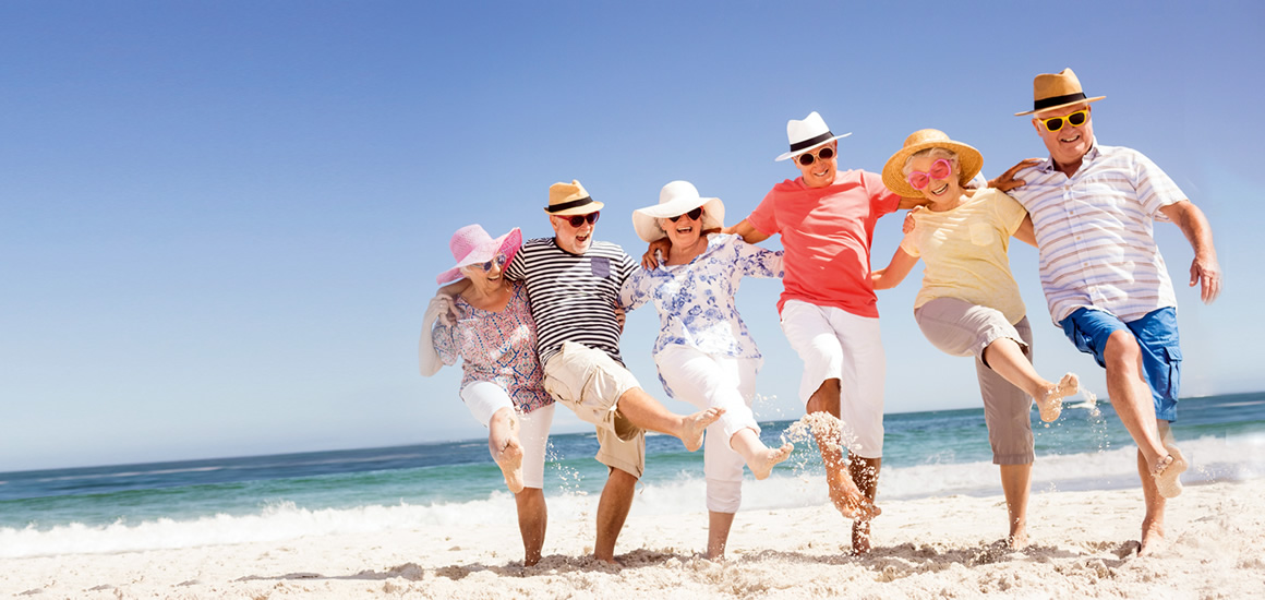 Eine Gruppe von Menschen tanzt fröhlich am Strand der Ostsee, alle tragen Sommerkleidung und Hüte.