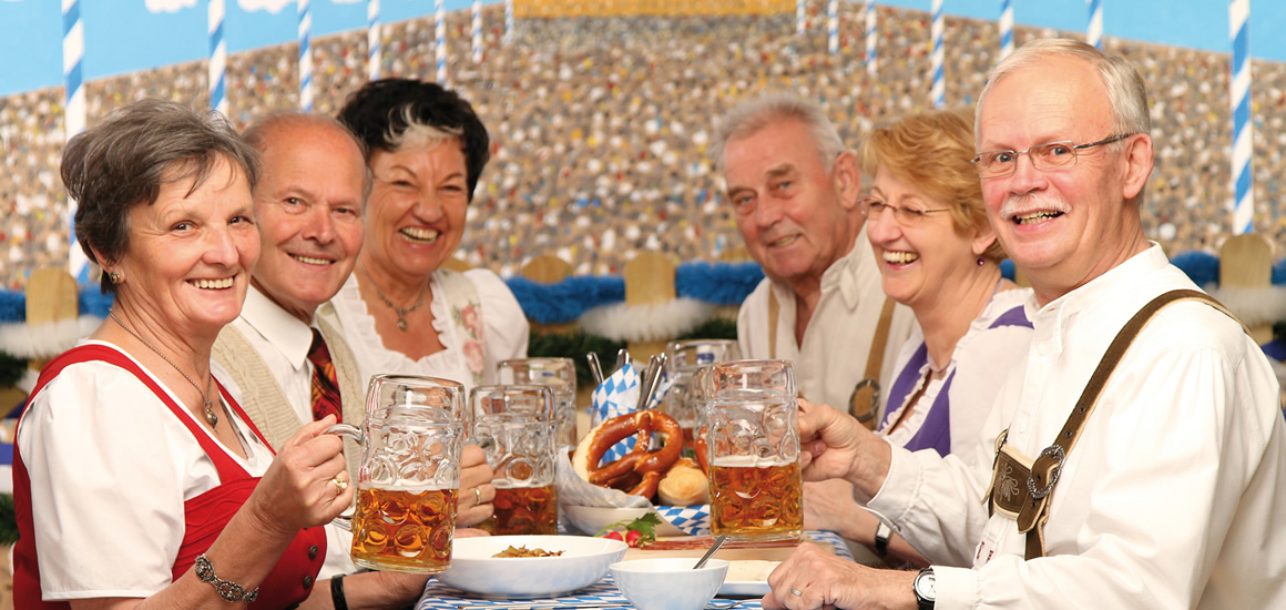 Eine Gruppe von Menschen in traditioneller bayerischer Kleidung sitzt an einem Tisch mit Bierkrügen und Brezeln, vermutlich auf einem Sommerfestival.