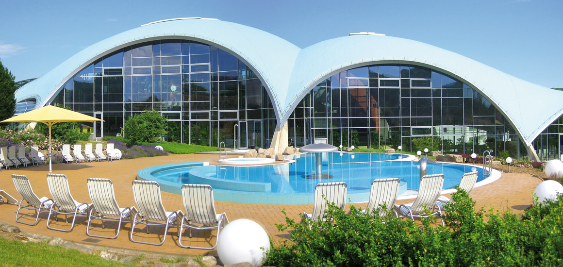 Außenansicht eines modernen Gebäudes mit geschwungenem Dach und großen Glasfronten, umgeben von einem Pool und Liegestühlen.