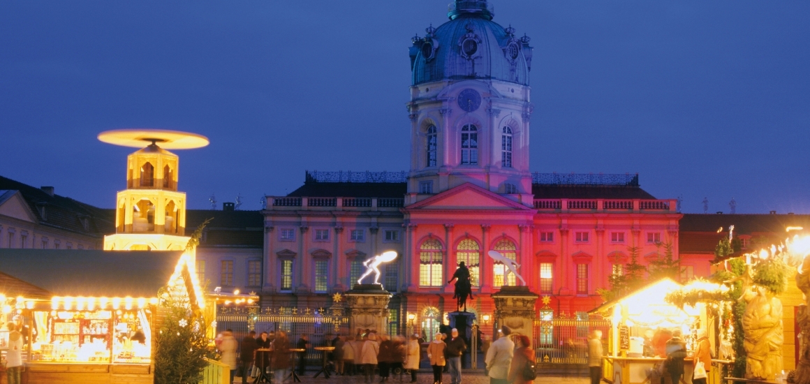 Weihnachtsmarkt vor dem Schloss Charlottenburg in Berlin, beleuchtet mit Ständen und einem großen Gebäude im Hintergrund.