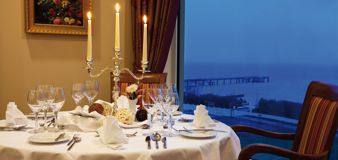 Ein elegant gedeckter Tisch in einem Restaurant mit Blick auf das Meer bei Dämmerung. Der Tisch ist mit einem Kerzenständer, Weingläsern und gefalteten Servietten dekoriert.