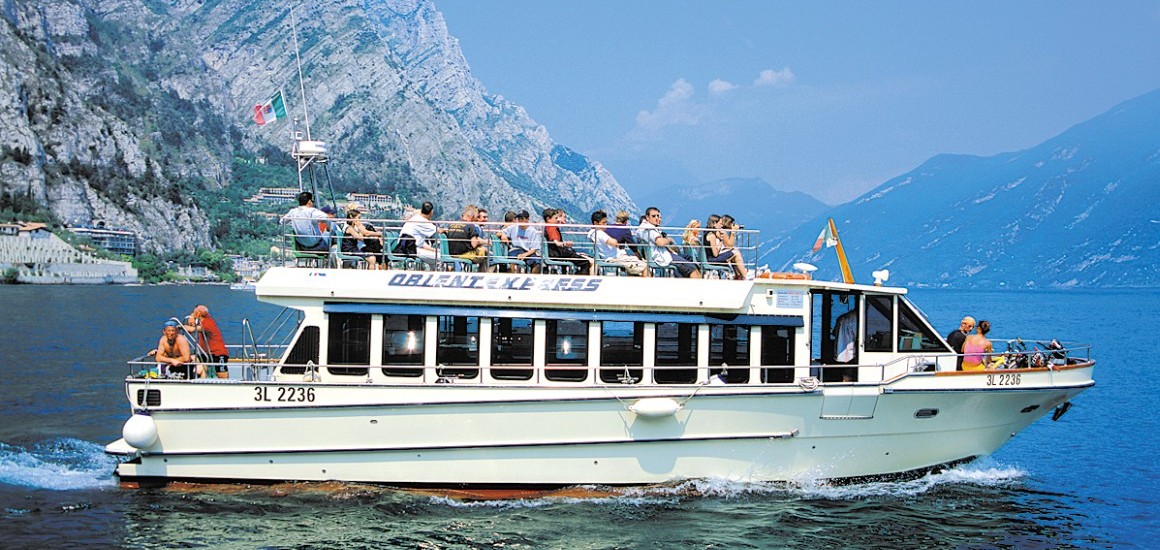 Ein Ausflugsboot auf dem Gardasee mit Passagieren an Bord, umgeben von Bergen und klarem Himmel.