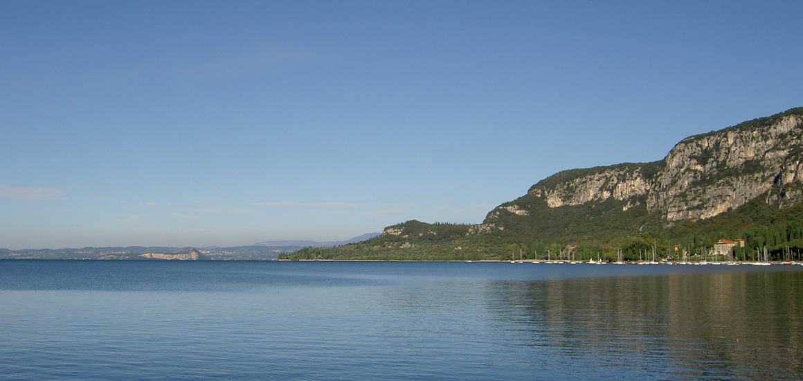 Foto vom Gardasee, ruhiges Wasser mit Bergen im Hintergrund unter klarem Himmel.