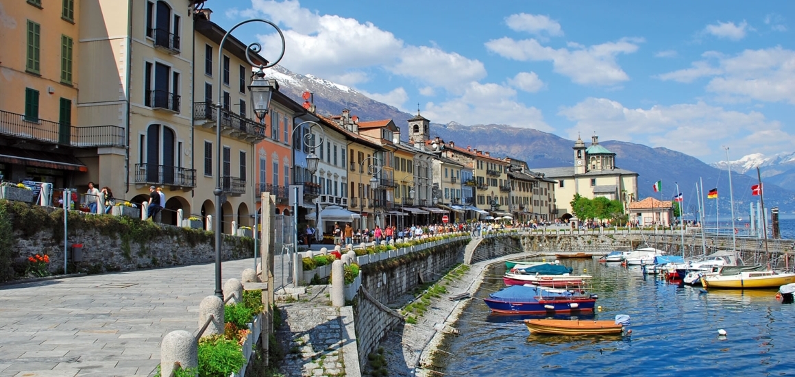 Uferpromenade in Cannobio, Italien, mit bunten Gebäuden, Booten im Wasser und Bergen im Hintergrund.